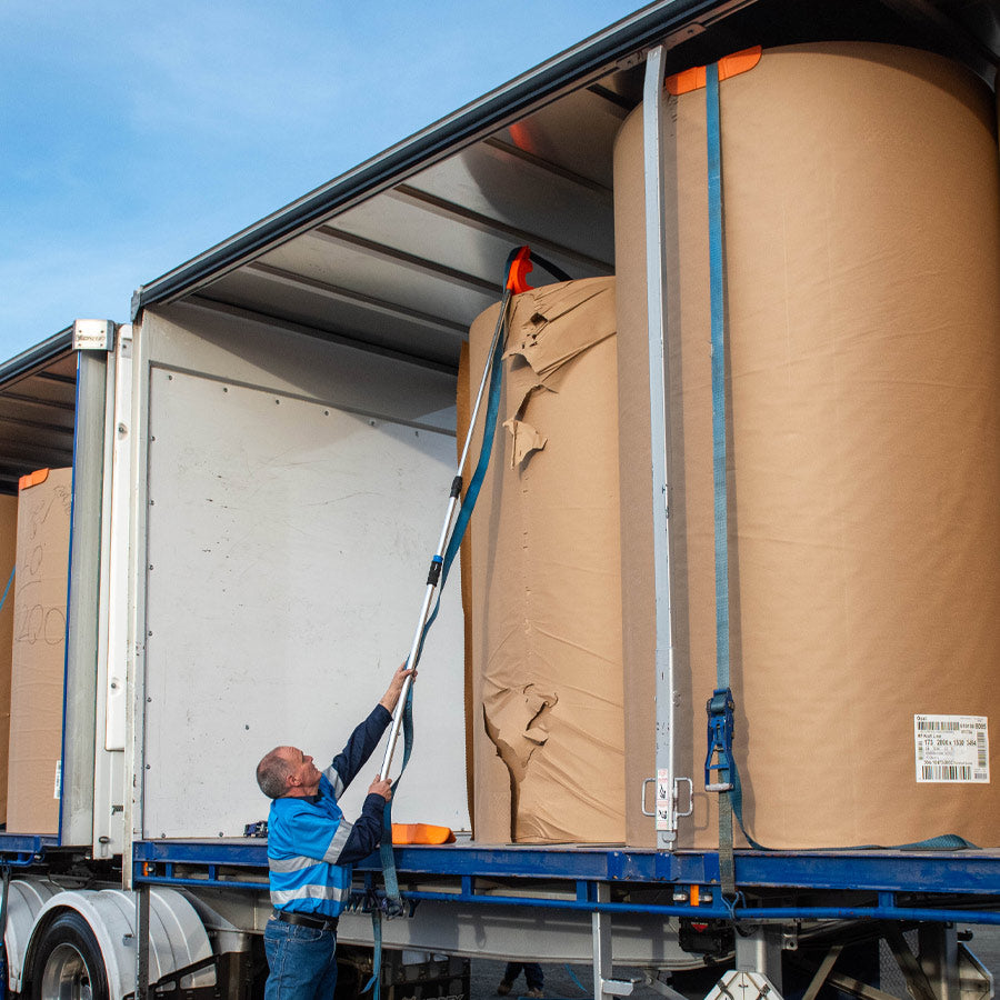 A man using an orange Strap Launcher tool on a pole to throw a tie-down strap over large cardboard rolls on a truck bed.