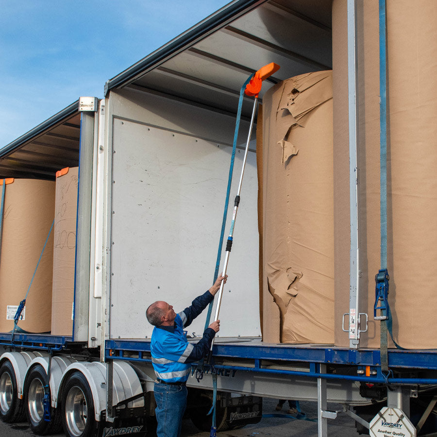 A man using an orange Strap Launcher tool on a pole to throw a tie-down strap over large cardboard rolls on a truck bed.