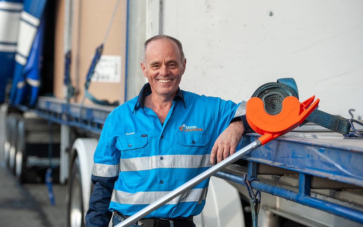 Owen Burns wearing blue hi-vis shirt, smiling and leaning against a truck bed, with an orange Strap Launcher tool ready to use.