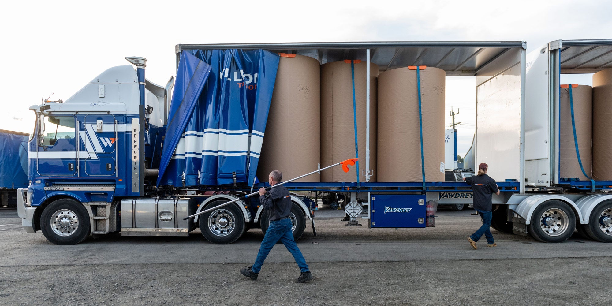 A man and woman securing straps after using an orange Strap Launcher tool on a pole to throw a tie-down strap over large cardboard rolls on a truck bed. 