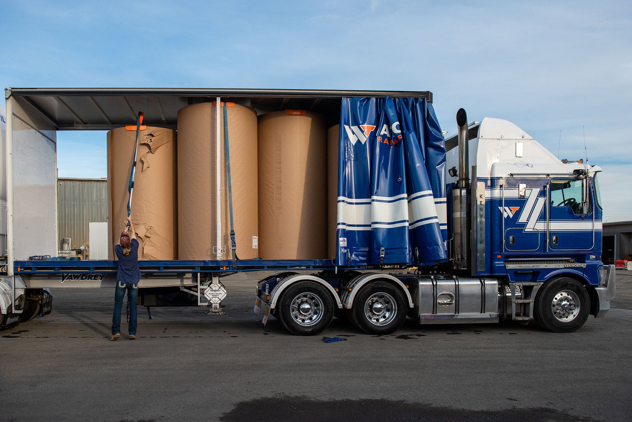 A woman using an orange Strap Launcher tool on a pole to throw a tie-down strap over large cardboard rolls on a truck bed.