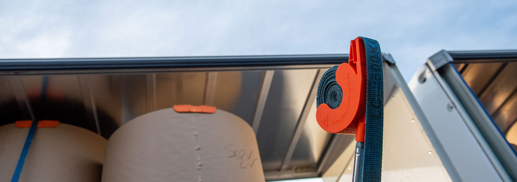 Orange Strap Launcher tool loaded with a blue tie-down strap and ready to throw over large cardboard rolls on a truck trailer.