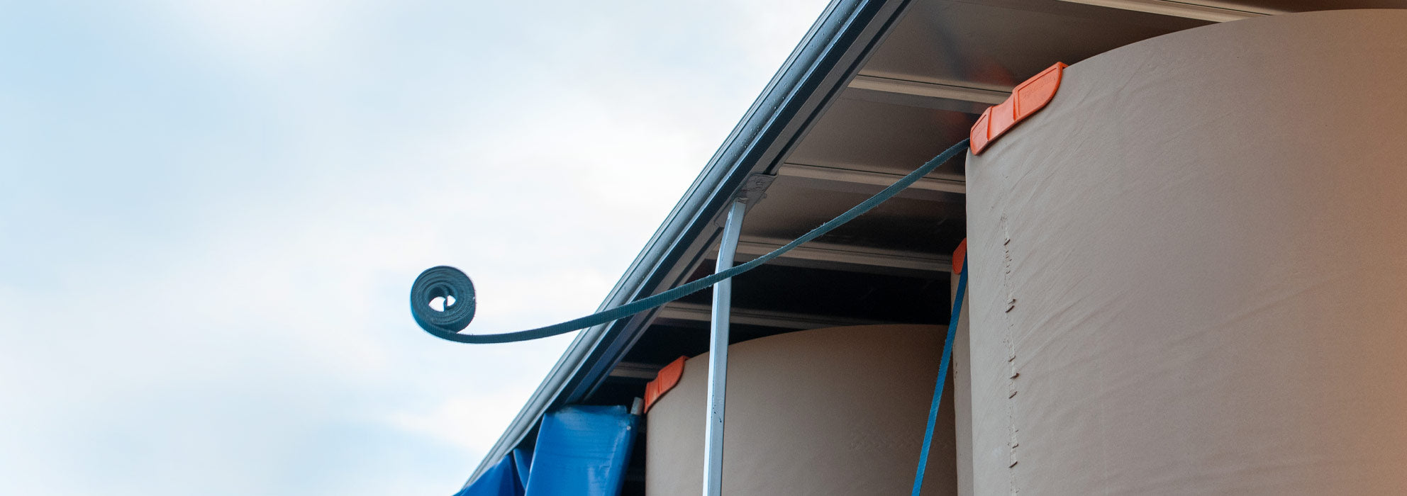 Looking up at a blue tie-down strap unrolling after being thrown over a tall item on a truck trailer.