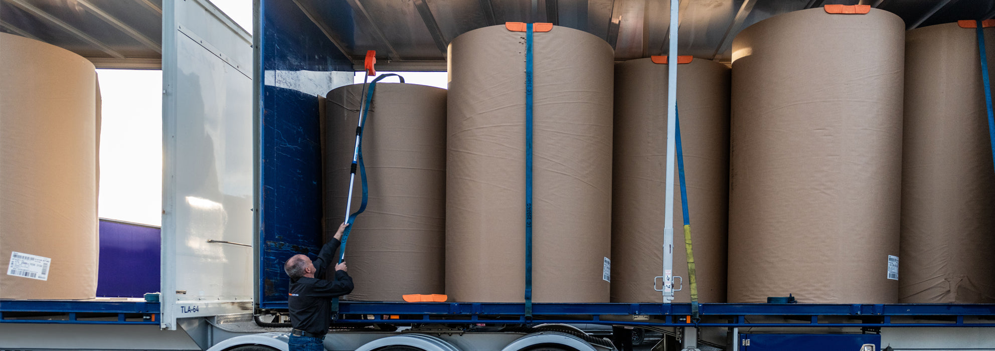 A man using an orange Strap Launcher tool on a pole to throw a tie-down strap over large cardboard rolls on a truck bed.