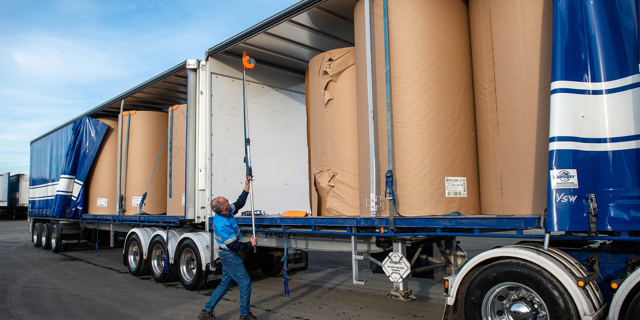 A man using an orange Strap Launcher tool on a pole to throw a tie-down strap over large cardboard rolls on a truck bed.