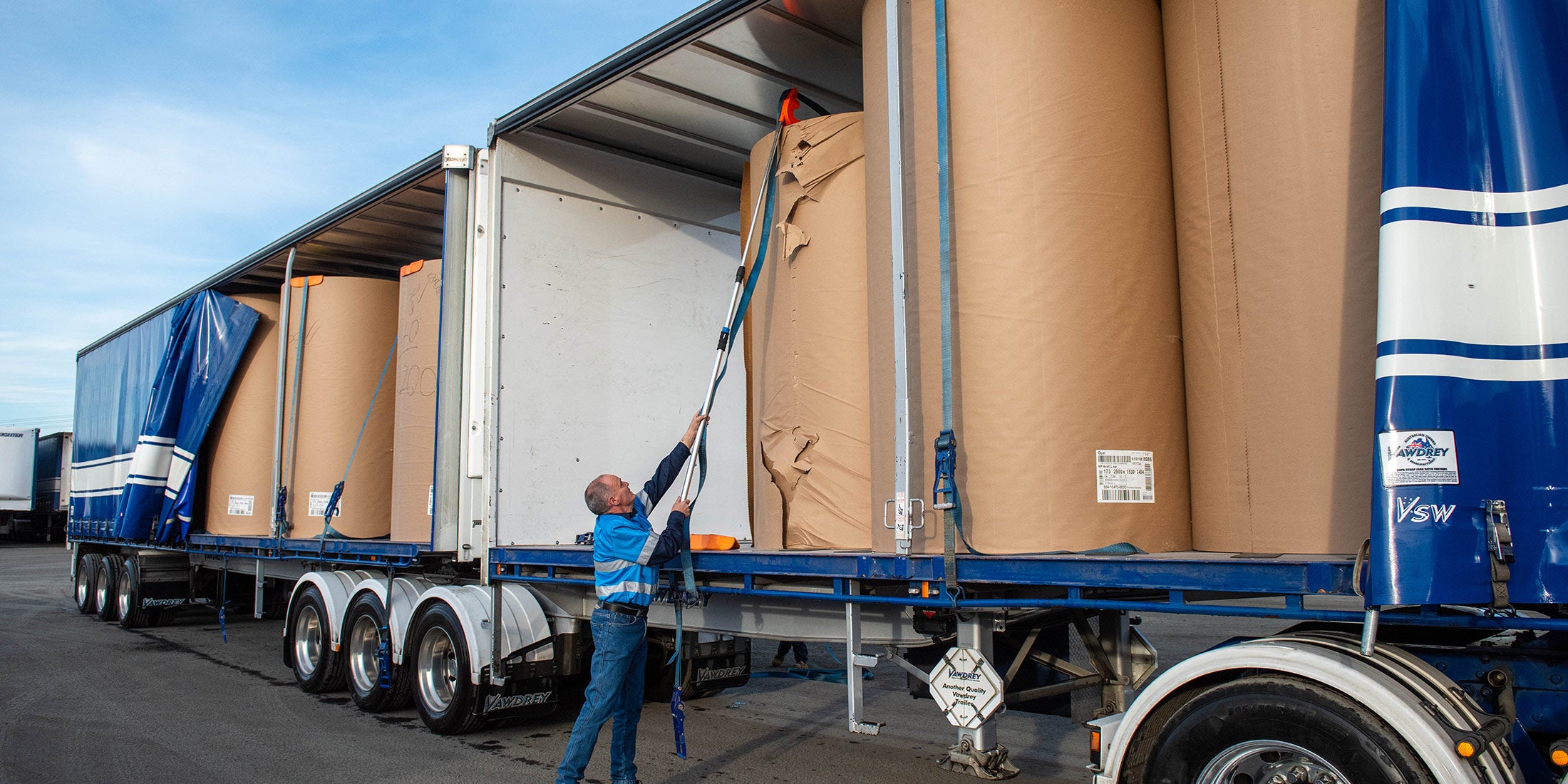 A man using an orange Strap Launcher tool on a pole to throw a tie-down strap over large cardboard rolls on a truck bed.