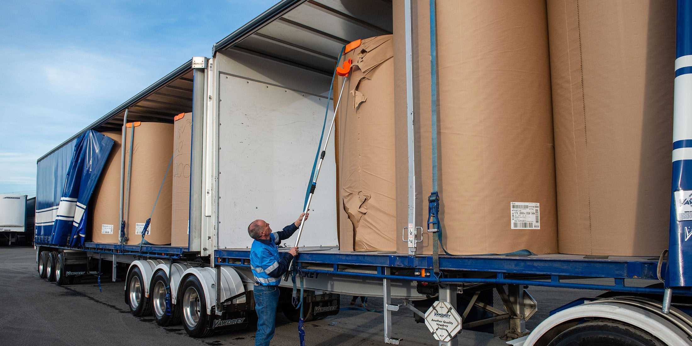 A man using an orange Strap Launcher tool on a pole to throw a tie-down strap over large cardboard rolls on a truck bed.